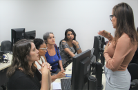 Foto de três mulheres sentadas em frente a computadores atentas ao que diz outra mulher de pé à sua frente.