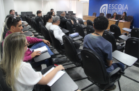 Foto panorâmica de plateia em auditório de paredes brancas, assistindo às falas de duas mulheres sentadas à frente, numa mesa de madeira, tendo atrás de si uma parede azul com a inscrição branca ESCOLA JUDICIAL.