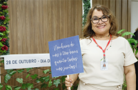 Foto de uma mulher de pele e cabelos claros, sorrindo para a câmera, usando óculos e vestido bege e segurando uma placa azul com o texto branco "NENHUM DE NÓS É TÃO BOM QUANTO TODOS NÓS JUNTOS!". Atrás dela, vê-se cenário decorado com flores.