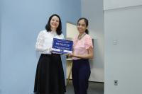 Foto de duas pessoas em pé, segurando uma placa. Ao fundo duas paredes, uma branca e outra azul.