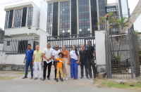 Foto de nove pessoas de pé e sorrindo para a câmera, e tendo ao fundo a fachada lateral do prédio-sede do TRT-16.