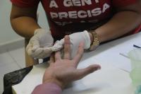 Foto mostra em detalhe as mãos de uma profissional de saúde fazendo a coleta sanguínea na mão de um paciente. 