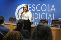 Foto mostra uma mulher em pé, diante de uma plateia. Ao fundo uma parede azul e a logo da Escola Judicial