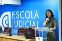 Foto mostra a juiza Carolina Burlamaqui posicionada no pulpito no auditório da EJUD. Ao fundo o Nome em letras brancas da Escola Judicial.