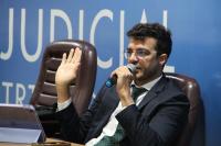 Foto mostra um homem branco usando um terno esculo e camisa azul claro, sentado em uma cadeira. Ele ergue a mão direita e com a mão esquerda segura um microfone. Ao fundo, um painel azul e o nome Escola Judicial.