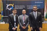 Arte feita com a foto de três homens, vestidos com terno. Eles estão em pé, posicionados em frente a um painel que tem informações sobre o Seminário Internacional de Precedentes Trabalhistas