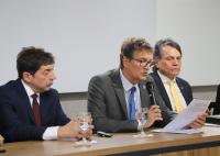 A imagem mostra três homens sentados a uma mesa. Todos vestem ternos. O homem ao centro fala ao microfone e segura um documento. Ele usa óculos e gravata azul. Há copos de água sobre a mesa. A parede ao fundo é branca e lisa.