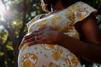 Foto parcial de uma mulher grávida, de pele escura e usando um vestido estampado de floral amarelo e branco, com a s mãos pousadas sobre a barriga. Atrás dela, árvores iluminadas por raios de sol.