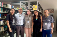 Foto mostra cinco pessoas em pé. Ao fundo prateleiras de alumínio com livros.