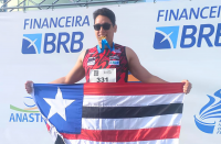 Foto mostra um homem com roupa esportiva. Ele está sobre o pódio de uma premiação. Ele segura a bandeira do maranhão. E tem na boca a medalha de ouro. Ao fundo um painel branco com detalhes em azul.