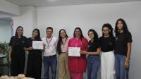 Foto mostra um homem e outras sete mulheres. Todos estão em pé no fundo de uma sala. Duas dessas mulheres seguram certificados. 