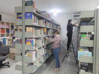 Foto mostra prateleiras com livros e duas pessoas entre as prateleiras. Uma delas, está em uma escada arrumando os livros e a outra em pé segura um livro.