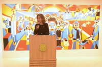 Foto mostra uma mulher em pé, atrás de um púlpito de madeira. Ao fundo uma pintura colorida retrata figuras que representam trabalhadores