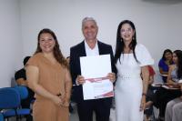 Foto mostra três pessoas em pé - são duas mulheres e um homem que segura um certificado. Ao fundo outras pessoas estão sentadas.