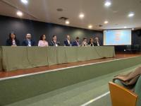 Foto mostra pessoas sentada atrás de uma bancada em um auditório.