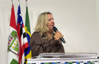 Mulher loira fala ao microfone, olhando para a direita e tendo, à esquerda, três bandeiras hasteadas.