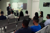 Foto mostra um homem, em pé usando terno escuro falando para pessoas que estão sentadas