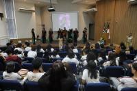 Foto mostra pessoas em apresentação no palco de um auditório. Outras pessoas estão sentadas nas cadeiras do auditório