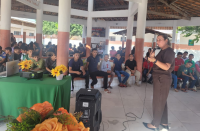 Foto mostra um mulher em pé, realizando uma palestra para jovens estudantes. 
