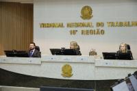 Foto mostra três pessoas sentadas. Ao fundo o brasão da Justiça do Trabalho e o nome Tribunal de Justiça do Trabalho da 16ª Região