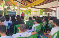 Foto mostra uma mulher fazendo uma palestra para jovens que estão sentandos. 