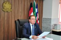 Um homem sentado segura uma folha de papel. Ao fundo, posicionado na parede o Brasão da Justiça e as bandeiras do Brasil, Maranhão e da Justiça do Trabalho