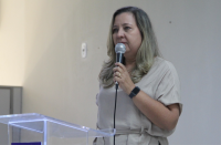 Foto de uma mulher branca, de cabelos claros, falando ao microfone, que segura com a mão esquerda, atrás de um púlpito transparente. Atrás dela, uma parede bege claro.