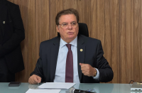 Foto de um homem sentado. Ele usa um terno escuro, camisa azul claro e uma gravata. 