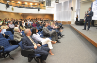 Foto mostra um homem em pé, realizando uma palestra. No lado direito várias pessoas estão sentadas acompanhando a apresentação do palestrante.
