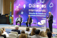 Foto mostra três homens usando terno escuro. Dois deles estão sentados e um em pé, falando ao microfone em um púlpito transparente. Atrás deles, telão de led exibe a identidade visual do evento. À frente deles, pessoas na plateia, sentadas em cadeiras brancas num auditório.