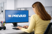 Foto de uma mulher clara de costas, usando blusa de mangas compridas amarela e cabelos longos. Ela está sentada a uma mesa, em frente a um computados, onde se lê na tela PREVJUD.