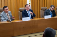 Foto de três homens claros, usando ternos e sentados em cadeiras marrons atrás de uma mesa de madeira. O do meio fala a um microfone, que segura com a mão esquerda.
