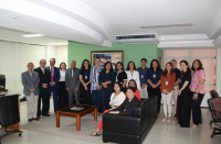 Foto mostra um grupo de pessoas, a maioria está em pé. Três delas estão sentadas em um sofá. 