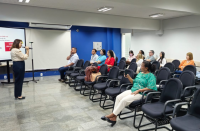 Foto mostra uma mulher realizando uma palestra. Tem algumas pessoas sentadas em cadeiras em um auditório