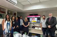 Foto mostra pessoas reunidas em uma sala. São homens e mulheres. Ambos estão em pe. Ao fundo uma muher aparece no monitor. Ela participa de maneira remota.