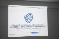 Arte com fundo branco, ao centro uma logomarca na cor azul formando um desenho de um coração, um escudo e um ouvido. Na parte inferior: Subcomitê de Prevenção e Enfrentamento do Assédio Moral, do Assédio Sexual e da Discriminação - 1º Grau