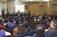 Foto mostra várioas pessoas sentadas em um auditório. Ao fundo, outras seis pessoas estão posicionadas no dispositivo de honra.