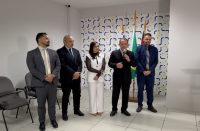Foto mostra cinco pessoas em pé; Ao fundo uma parede branca com azulejos