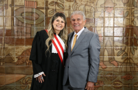 Foto mostra um homem e uma mulher. Ambos estão em pé. Ao fundo uma parede com azulejos marrons.