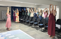 Foto mostra várioas mulheres, dentro de um auditório. Todas estão realizando movimento laboral e estão com os braços erguidos.