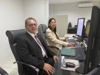 Foto mostra um homem e uma mulher sentados lado a lado, ambos estão diante de monitores de computador.