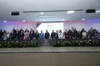 Foto mostra um palco. Sobre o mesmo, várias pessoas em pé ocupando o dispositivo de honra.