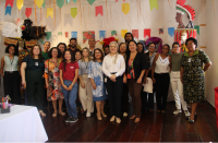 Foto mostra várias pessoas reunidas em uma sala. Ao fundo uma parede branca e decoração com bandeirinhas coloridas. 