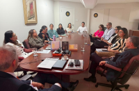 Foto mostra várias pessoas sentadas ao lado de uma grande mesa de reunião.
