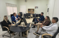 Foto mostra pessoas sentadas em volta de uma mesa de reuniões. No centro da mesa, computadores.