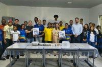 Foto mostra várias pessoas em pé. Todos estão posicionados atrás de uma grande mesa. Duas pessoas seguram um cartaz com o nome PID