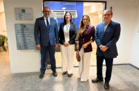 Foto mostra quatro pessoa em pe, um ao lado da outra. ao fundo a imagem da sala do CEJUSC 2º Grau