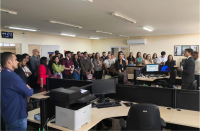 Foto mostra vários pessoas reunidas em pé, dentro de uma grande sala. 