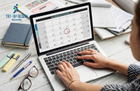 Foto de uma mesa de trabalho com mãos femininas no teclado de um notebook, em cuja tela aparece um calendário.