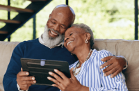 Foto de um homem e mulher negros e idosos abraçados e sorrindo, olhando a tela de um tablet, sentados em um sofá.